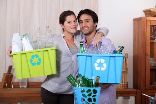 A young couple standing indoors in a home environment, holding two plastic recycling bins filled with empty glass bottles. The woman on the left is wearing a light grey top and has dark hair tied back, while the man on the right is dressed in a light purple shirt and has dark, curly hair. The green bin with a recycling symbol contains clear glass bottles, and the blue bin with a recycling logo holds additional bottles, some with green tint and others clear. A blue plastic container with a leaf pattern is also visible, filled with more glass bottles. Behind them, there is a wooden cabinet and a neutral-colored wall, indicating a kitchen or living space. The lighting appears natural, emphasizing the textures of the bottles and the smooth surfaces of the bins. The scene reflects an active waste recycling effort typical of a household working towards rubbish management services, such as those provided by Rotherhithe House Clearance in the local Rotherhithe area.