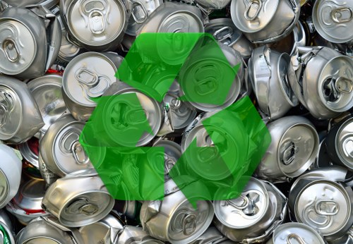 A close-up photograph of numerous stacked aluminium beverage cans, primarily silver with some showing red and white branding elements, featuring pull-tab tops and smooth, curved surfaces. The cans vary slightly in size and orientation, with some lying flat and others tilted or leaning against each other, creating a dense pile. The background consists entirely of additional cans, filling the frame. Superimposed over the cans is a prominent green recycling symbol, comprising three chasing arrows forming a triangle, indicating waste recycling. The overall scene is well-lit, highlighting the metallic sheen and textured surfaces of the cans, suggesting an environment related to waste collection or recycling efforts, possibly in the context of rubbish removal services such as those offered by Rotherhithe House Clearance, focused on recycling and waste management in the Rotherhithe area.