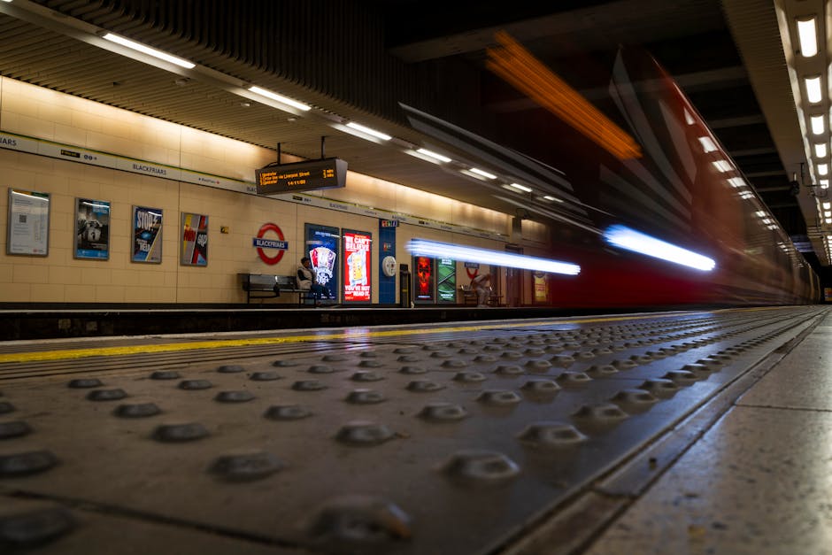 A London Underground station platform with beige tiled walls and wooden ceiling panels. Advertisements and station signage are visible along the wall, including a red and white sign for the 'Blackfria